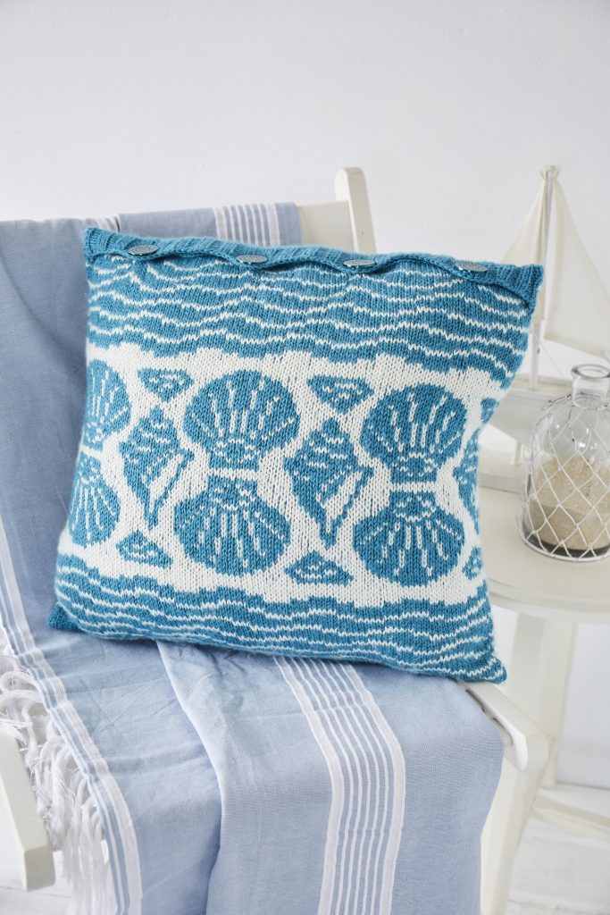 Knitted cushion with blue and white shell designs sits on deck chair in white room