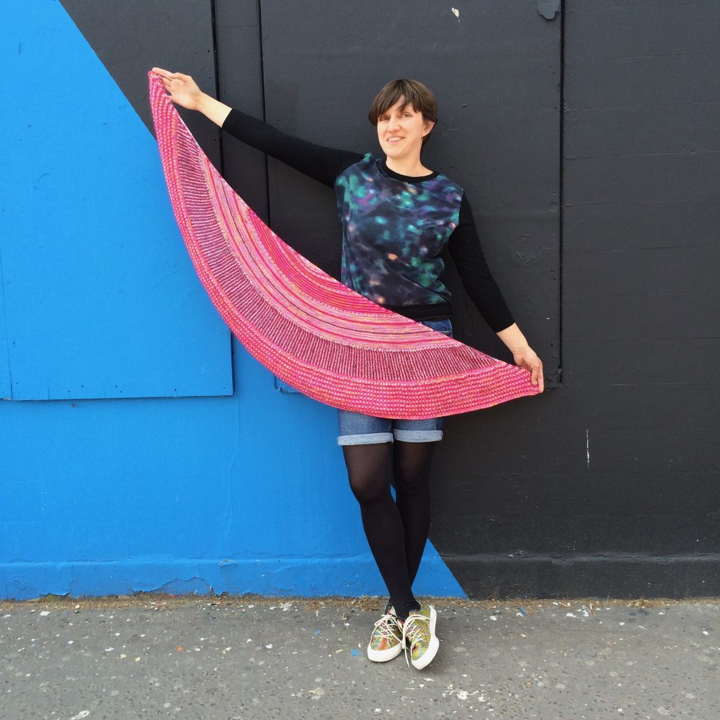 Photo of Amelia holding up a pink crescent shawl. She stands in front of a wall which is painted blue and grey.